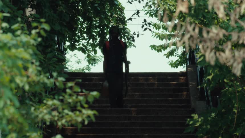 A young Black man with dreadlocks and a red backpack walks down a shady, tree-lined staircase carrying a skateboard
