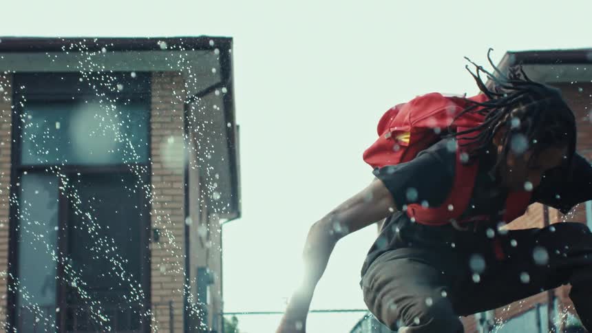 Slow motion shot of a young man with dreadlocks and a red backpack playfully jumping through a backyard sprinkler