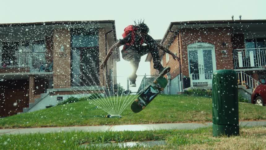 A skateboarder performs a trick over a lawn sprinkler in cinematic slow motion, landing on a hydrant in a sunny suburban neighborhood