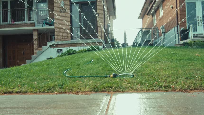 A lawn sprinkler oscillates back and forth, watering a green grassy yard in a suburban neighborhood on a summer day
