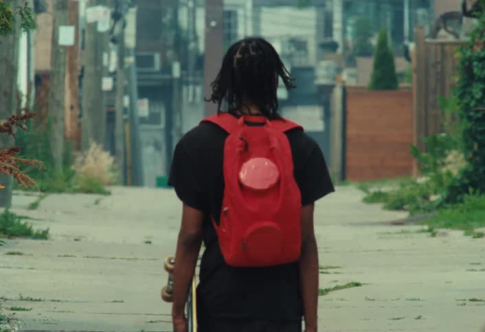 A young Black man with a red backpack walks away from the camera in slow motion down a gritty city alley, carrying his skateboard