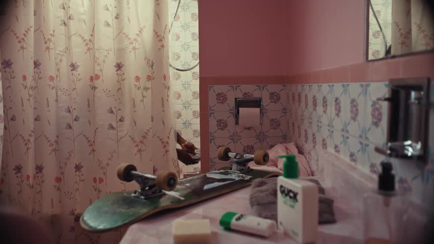 A person showers behind a floral curtain in a retro pink bathroom with a skateboard on the counter
