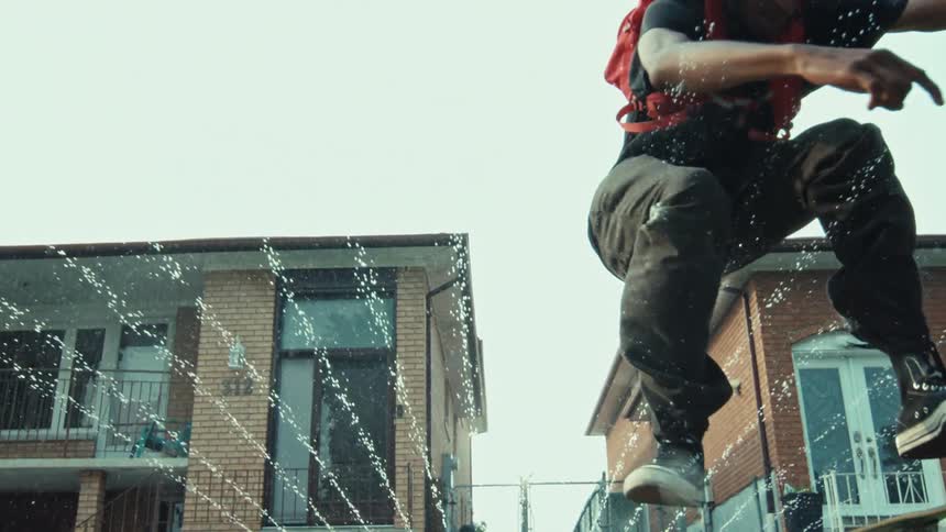 A young man with a backpack on a skateboard leaps over a fan of water from a sprinkler in a suburban yard in slow motion