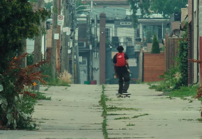A skateboarder with a red backpack performs a trick while riding down a long, gritty city alleyway