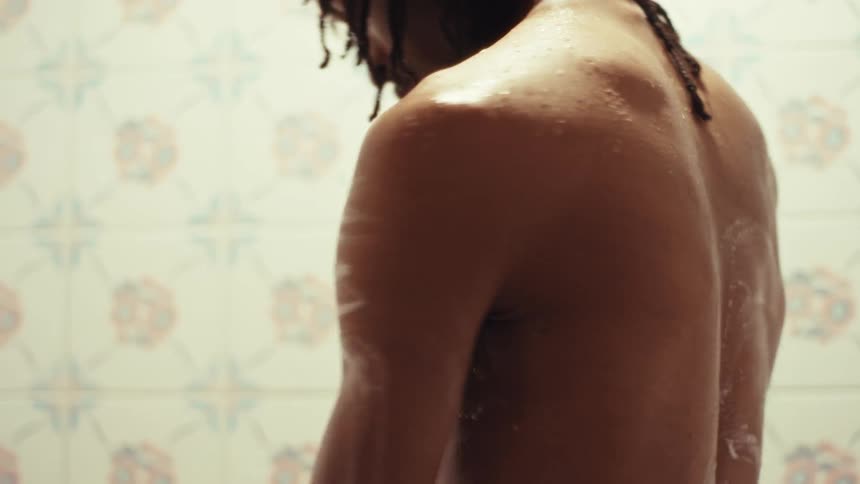 A muscular man with dreadlocks lathers his body with soap in a slow-motion shower