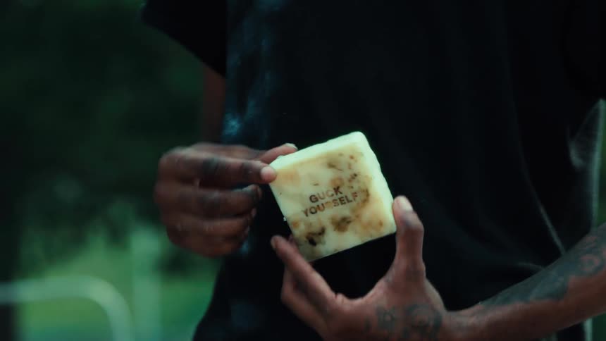 A person with tattooed arms holds up a handmade bar of soap with the words 'Guck Yourself' stamped on it