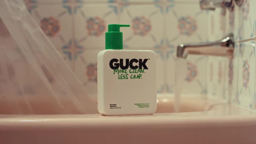 A floral shower curtain is pulled back to reveal a bottle of body wash on the edge of a bathtub with the faucet running