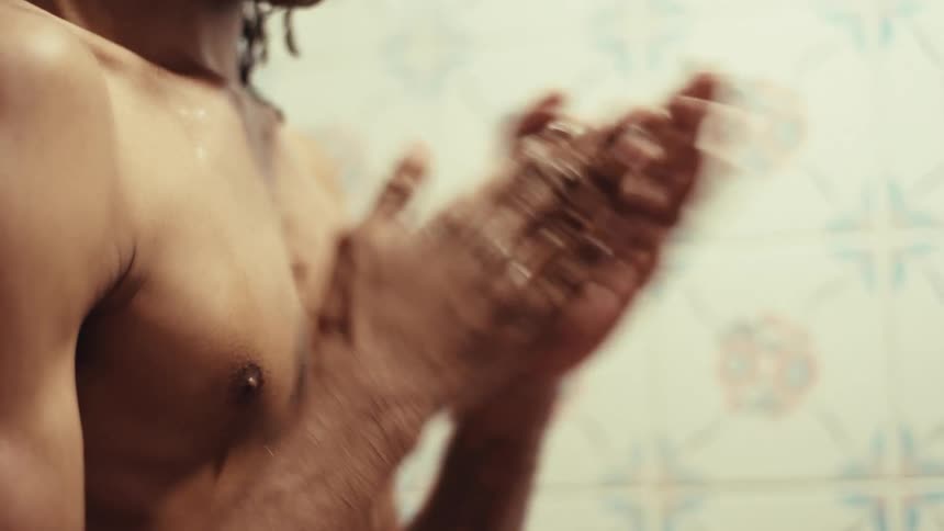 Close-up of a shirtless man with dreadlocks lathering soap on his muscular chest and arms in a tiled shower