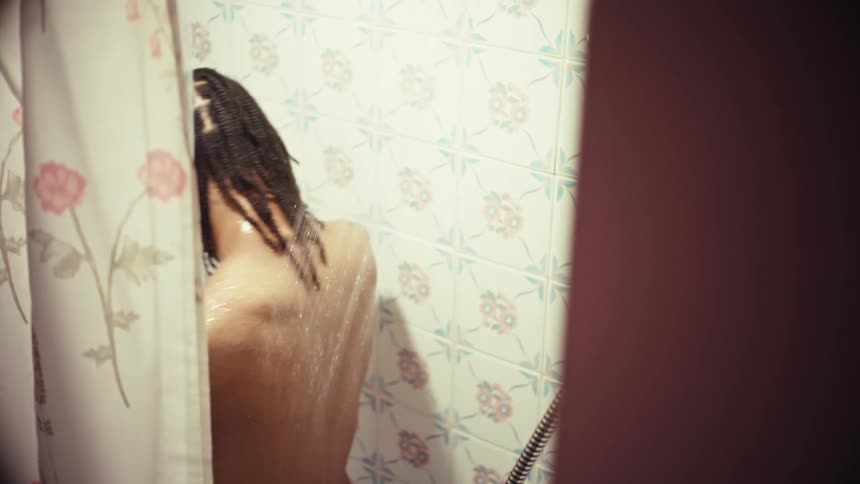 A partially obscured shot shows a man with dreadlocks taking a refreshing shower in a tiled bathroom