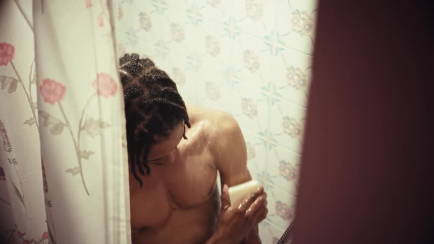 A muscular man with dreadlocks washes his body with a bar of soap while showering in a vintage-style bathroom