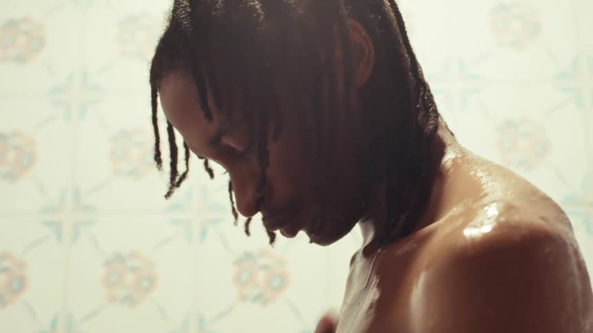 An athletic man with dreads enjoys a relaxing and slow-motion shower in a tiled bathroom