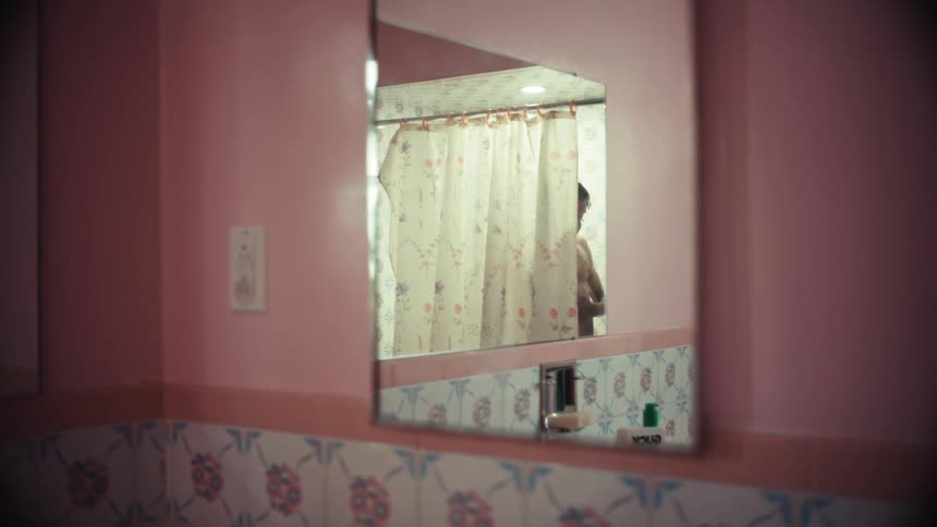 A person's silhouette is reflected in a bathroom mirror as they shower behind a floral curtain in a pink tiled room