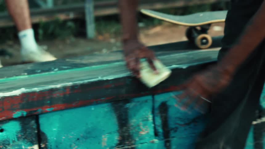 A skateboarder with tattooed arms applies wax to a graffiti-covered ledge at a skatepark