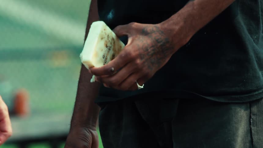 A close-up of a tattooed man's hand holding a block of skateboard wax at a park
