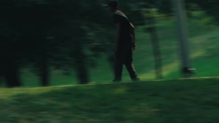 A young man skateboards down a winding path in a park and runs across a field in slow motion