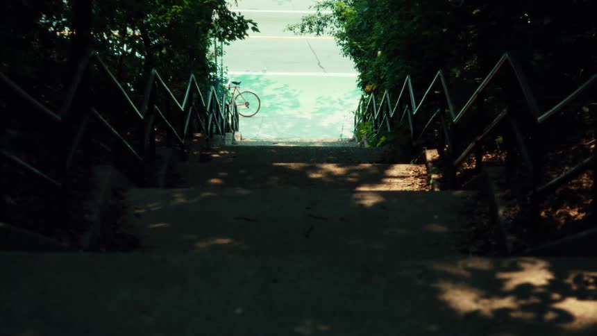 A high-angle view from the top of a shaded staircase shows a skateboarder riding along a bright green bike lane