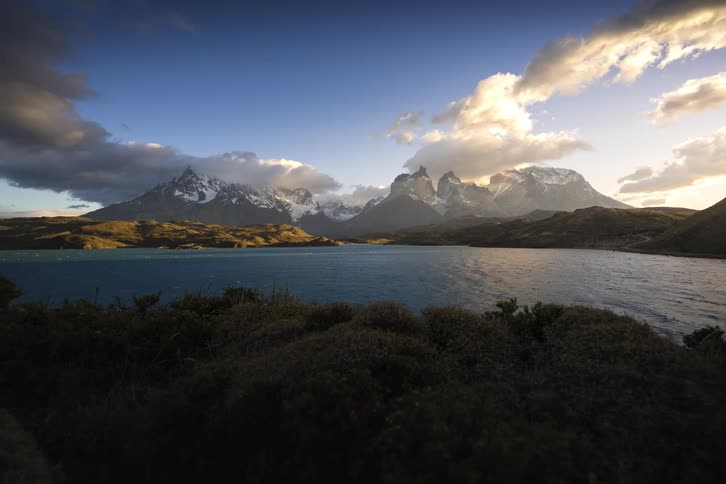 A dramatic time-lapse of a sunrise over the iconic Cuernos del Paine mountains, with clouds swirling and golden light hitting the peaks above Lake Pehoé