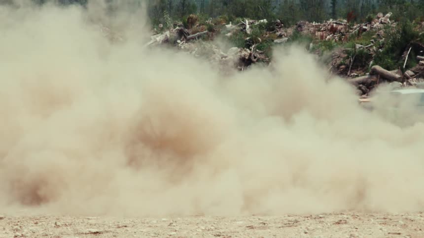 A black car kicks up a massive cloud of dust while doing donuts in a junkyard on a sunny day