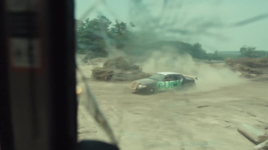 POV from a construction vehicle with a cracked windshield watches a demolition derby car drifting in a dusty junkyard