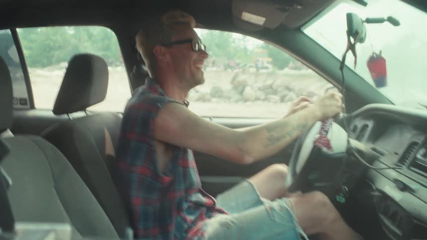 A smiling man in a sleeveless plaid shirt joyfully drives his car in an off-road setting, fist-pumping in excitement