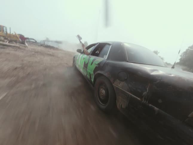 A person with a weapon leans out of a fast-moving, beat-up car kicking up dust on a dirt road in a seamless loop