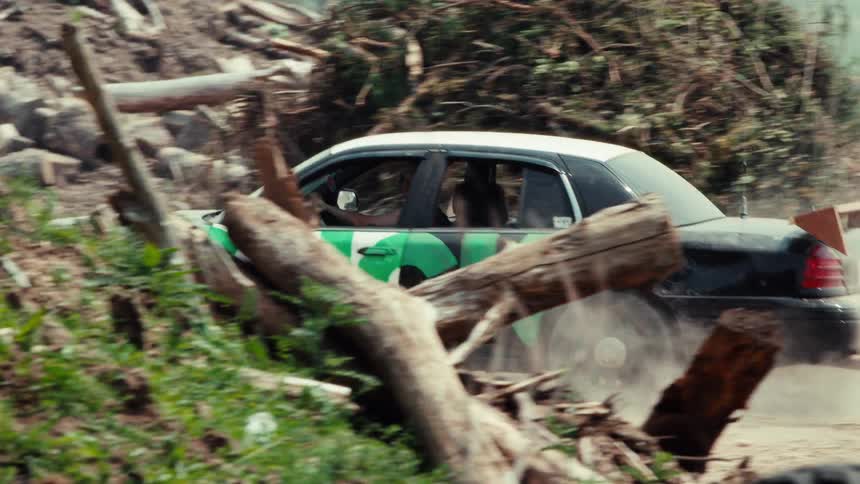 A customized black and white race car speeds down a dirt track in a rural junkyard, kicking up dust as it passes