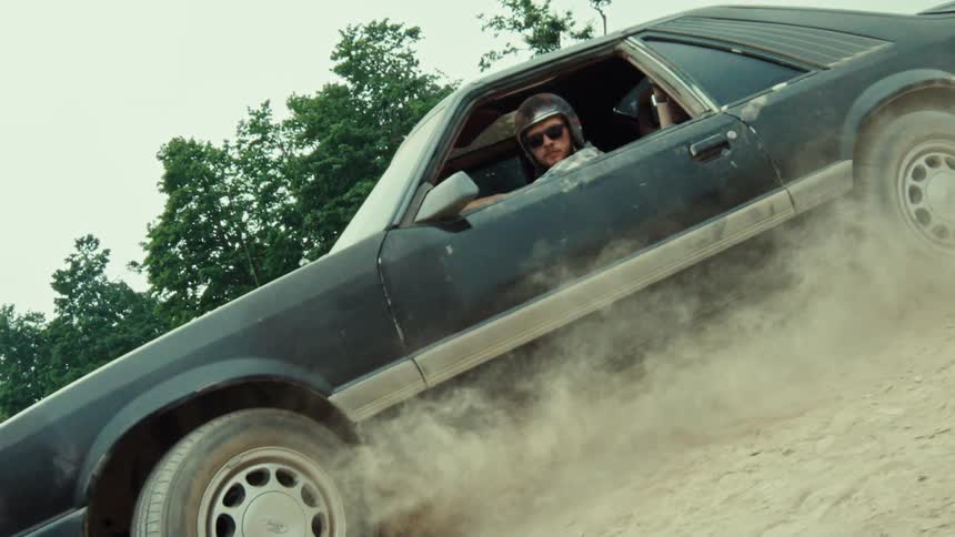 Slow motion shot of a man in a helmet doing a burnout in a vintage black muscle car on a dirt road, kicking up a cloud of dust