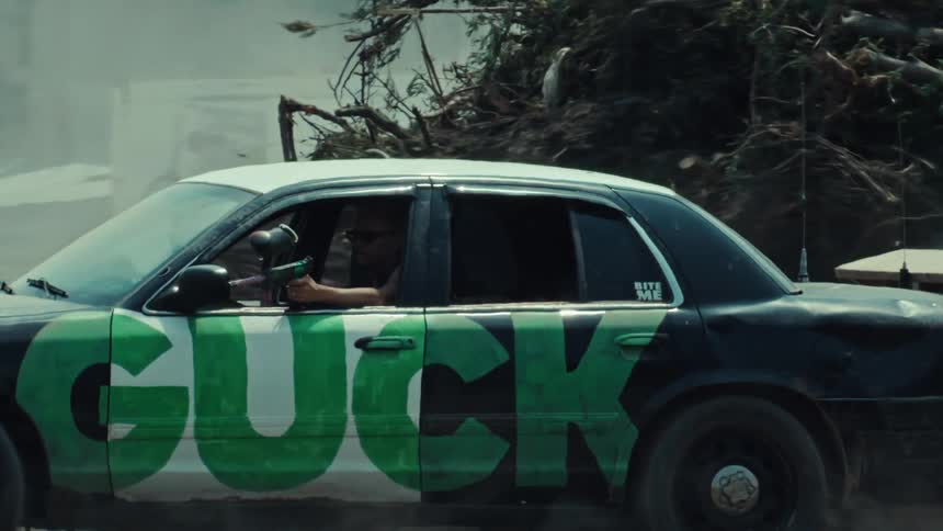 A man in slow motion aims a paintball gun from the window of a graffiti-covered police car in a junkyard