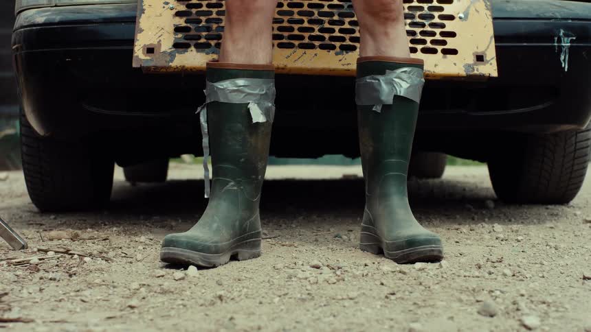 A man duct tapes his dirty green rubber boots to his bare legs in front of an old car
