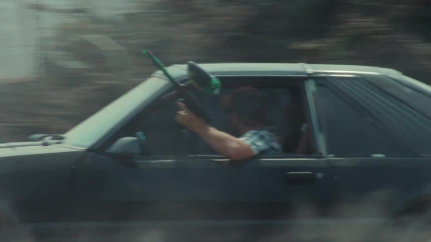 Slow motion shot of a man firing a paintball gun from a speeding vintage black car on a dusty road