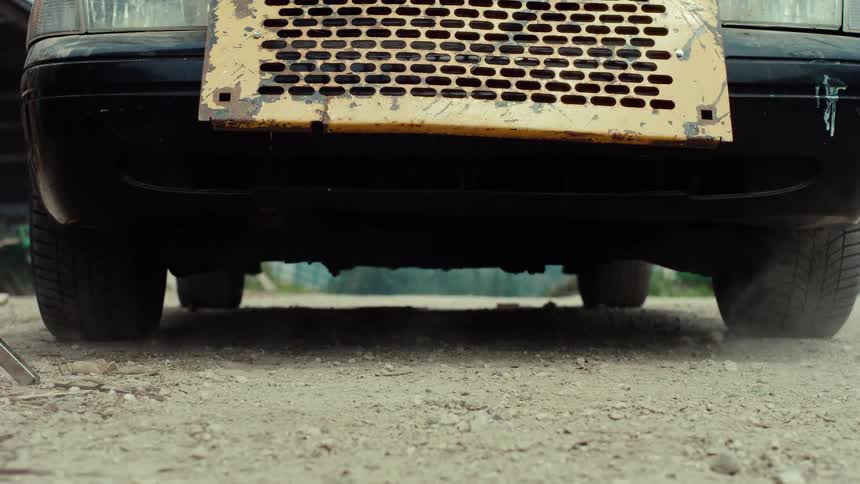 A close-up of a person wearing duct-taped rubber boots shuffling their feet and kicking up dust before walking away from an old car