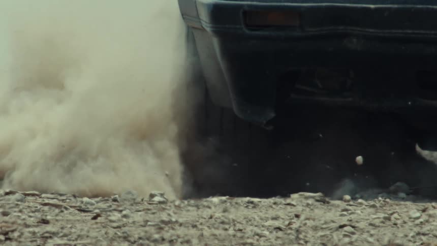 A close-up of a car tire spinning out on a dirt road, kicking up a cloud of dust and rocks in super slow motion