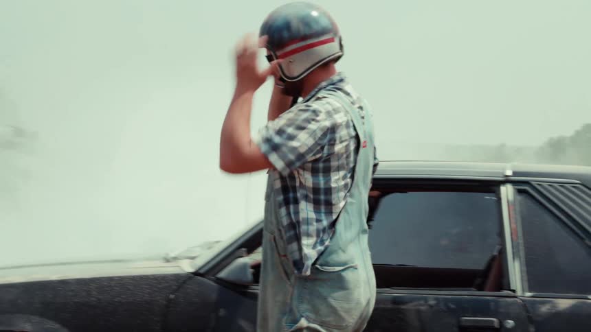 A man in overalls and a helmet gets out of his smoking black sports car, removes his helmet in frustration, and walks away in slow motion