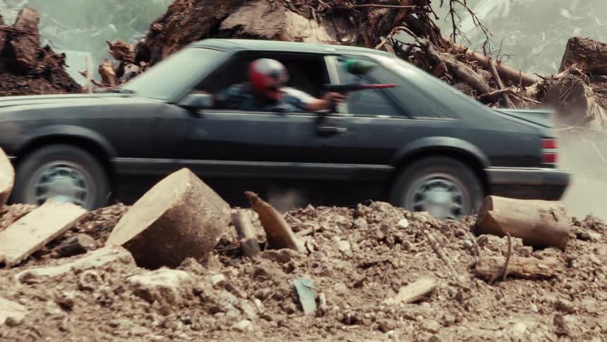 A man in a helmet shoots a paintball gun from a speeding Mustang during a chaotic chase in a dusty wasteland