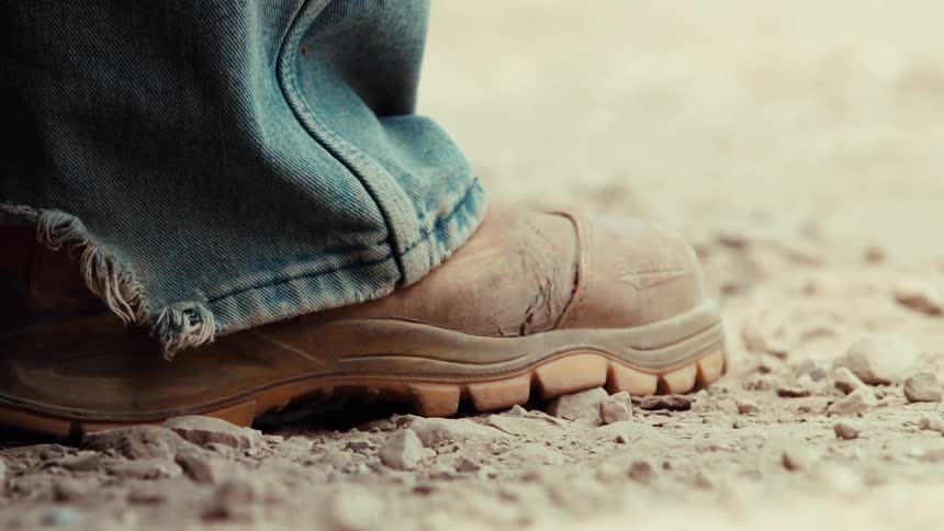 Close-up of a worker's worn-out, dusty boot tapping on the gravel ground before walking away