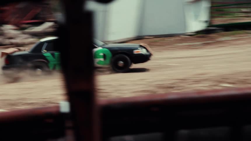 A handheld tracking shot follows a modified sedan with green graffiti as it speeds around a dusty dirt track in a junkyard