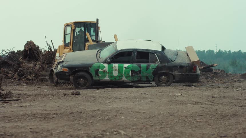 A yellow bulldozer slowly crushes a black sedan with green graffiti spray-painted on the side in a dirt junkyard