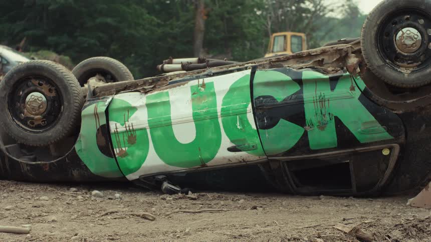 A wrecked car with the word 'cuck' spray-painted in green on its side lies overturned in a junkyard