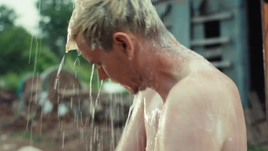 A shirtless man with bleached blonde hair and tattoos takes a shower outdoors, standing barefoot in the mud