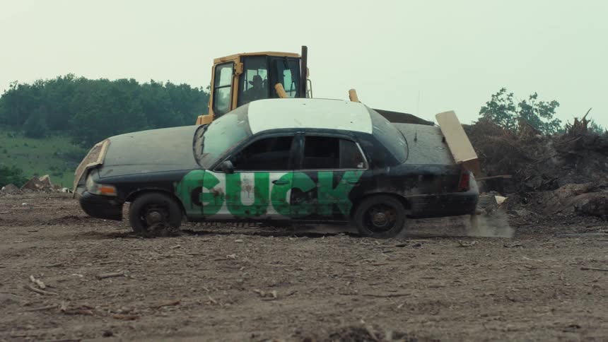 A bulldozer pushes a graffitied car through a junkyard in slow motion