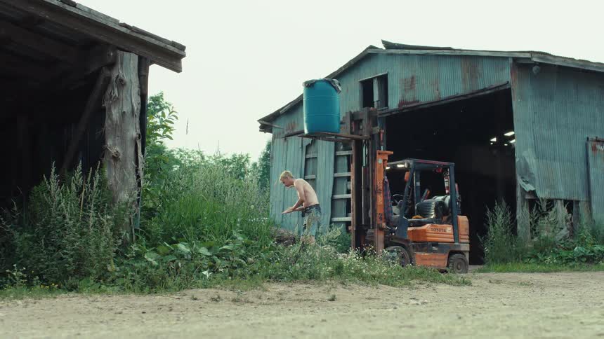 A shirtless worker chops wood with an axe next to a forklift in a rustic, rural setting