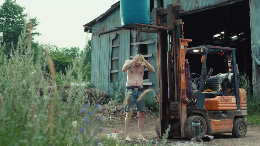 A resourceful man takes an improvised outdoor shower using a forklift to hold a barrel of water in front of a rustic barn