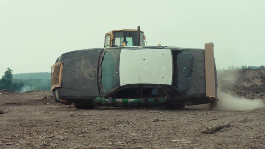 A powerful bulldozer slowly flips a derelict sedan with graffiti on its side in a dusty, barren landscape