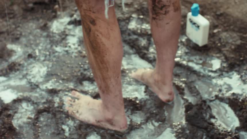 A barefoot person in denim shorts washes thick mud off their legs with a special cleaner while standing in a muddy puddle