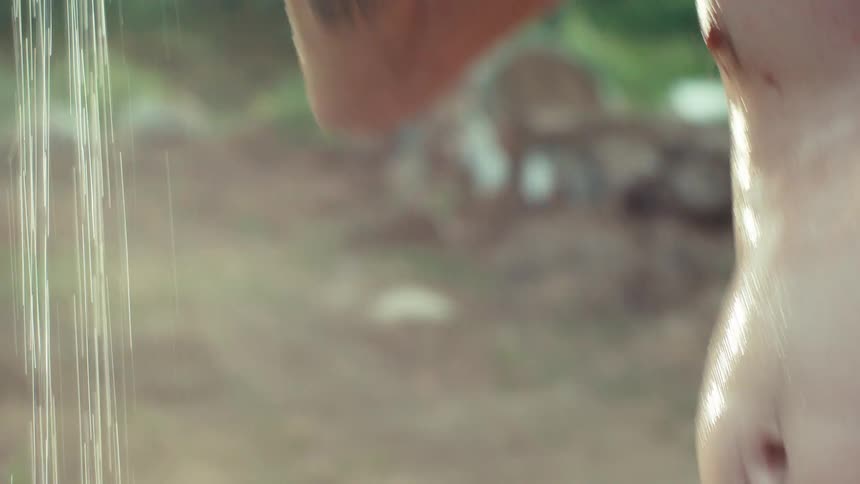 Slow motion close-up of a tattooed, shirtless man washing the dirt and grime off his hands and hair under an outdoor shower