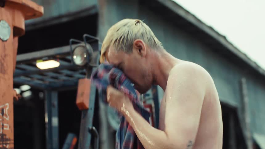 A shirtless tattooed man with bleached blonde hair washes himself with his wet plaid shirt in the rain at a rural worksite