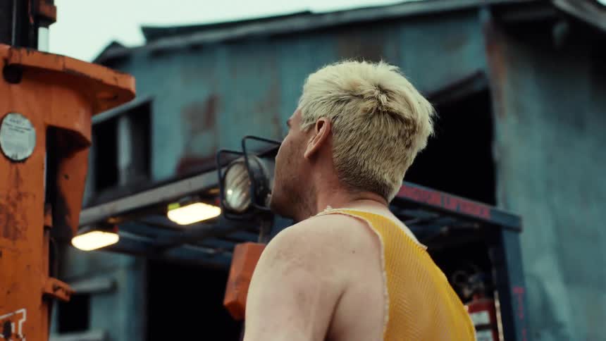 A barefoot man with muddy legs and feet takes off his dirty plaid shirt and mesh tank top next to a forklift