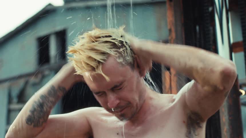A shirtless, tattooed man with bleached blonde hair washes himself outdoors in a muddy, industrial setting in slow motion