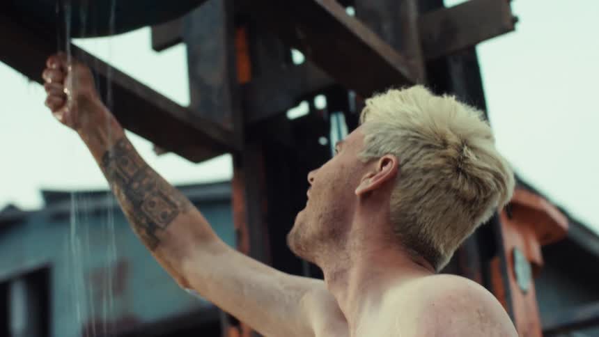 A shirtless man with tattoos and blonde hair takes a refreshing DIY shower from a water-filled barrel lifted by a forklift