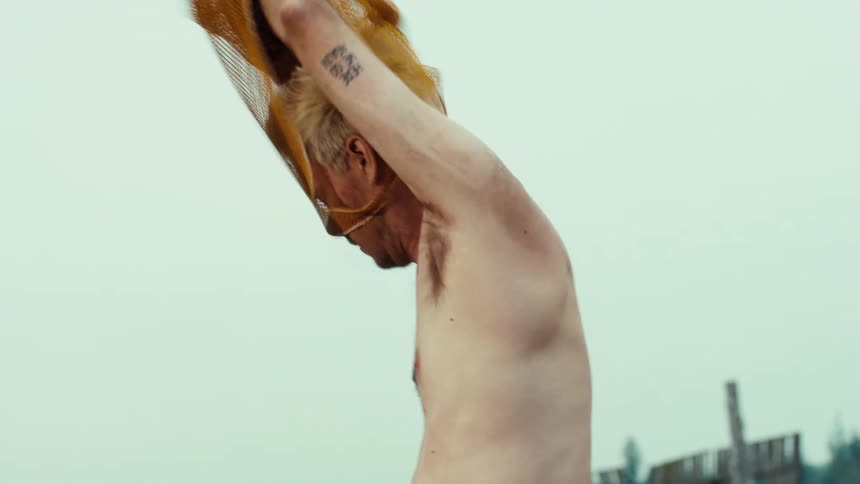 Bleached-haired man removes mesh tank top beside rusty industrial machinery in a dusty yard